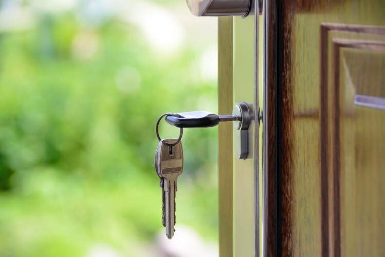 A key is inserted into the keyhole of a door with a keychain and two other keys attached, both keys hanging down. The door is slightly ajar, and a blurred, green outdoor scene is visible in the background.