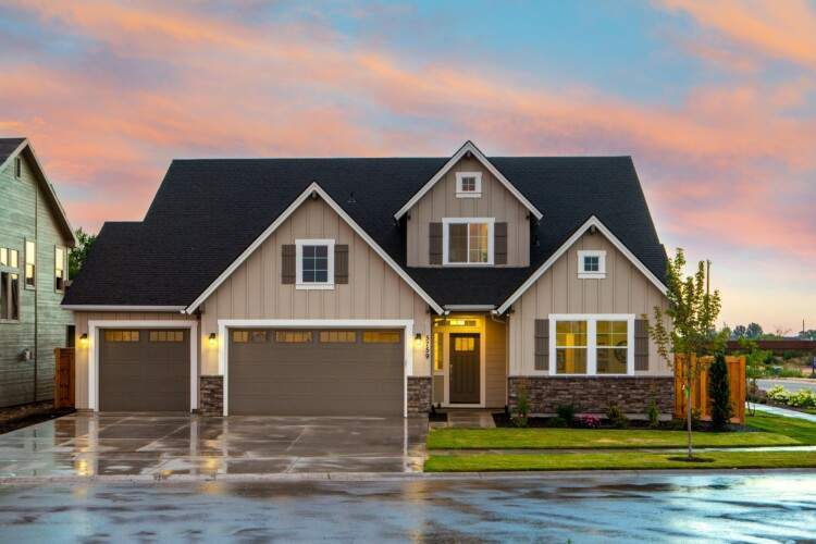 A modern house with beige siding, a black roof, and three garage doors. The front yard features a small lawn and a young tree. The sky is pink and cloudy, suggesting either sunrise or sunset.