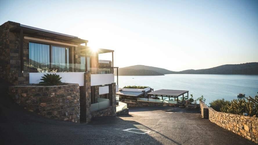 Modern stone house with large glass windows overlooking a serene lake at sunrise. The property features a patio area and lush surrounding hills with a clear road leading to the house.