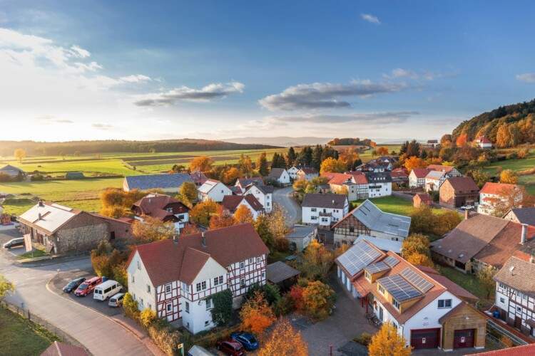Aerial view of a small village with red-roofed houses, surrounded by green fields and trees with autumn foliage under a partly cloudy sky.