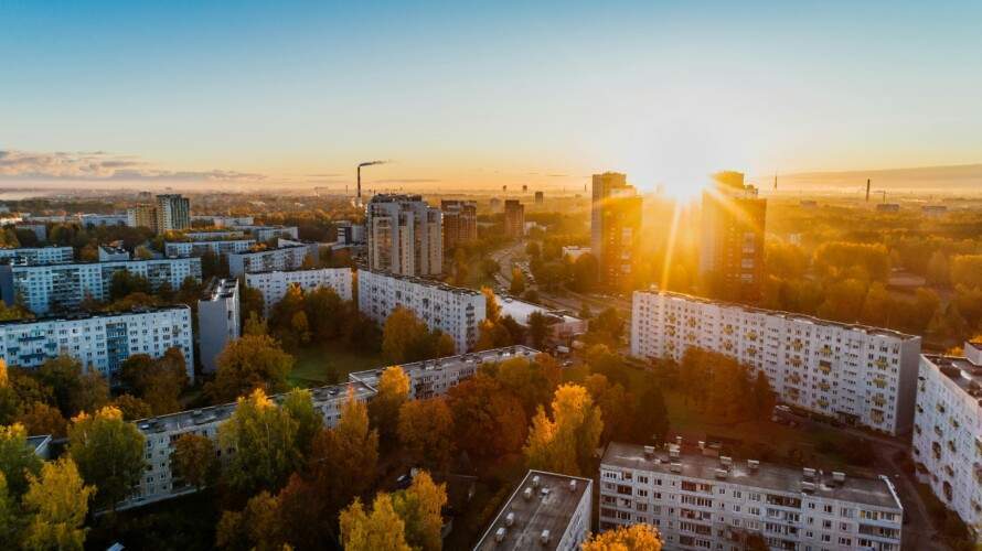 Aerial view of a cityscape with residential buildings, trees with autumn foliage, and the sun setting behind distant high-rise structures.