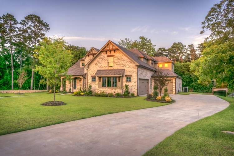 Large stone house with a well-maintained lawn, surrounded by trees. The driveway is curved and leads to an attached garage.