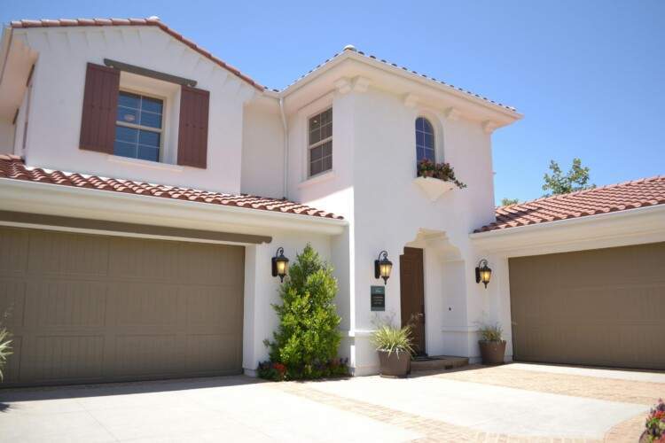 Two-story white house with brown roof tiles, twin garage doors, a small balcony with plants, and outdoor lantern-style lights above the garage.