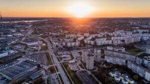 Aerial view of a cityscape during sunset, with residential buildings, roads, and parking lots in the foreground, and the sun setting on the horizon.