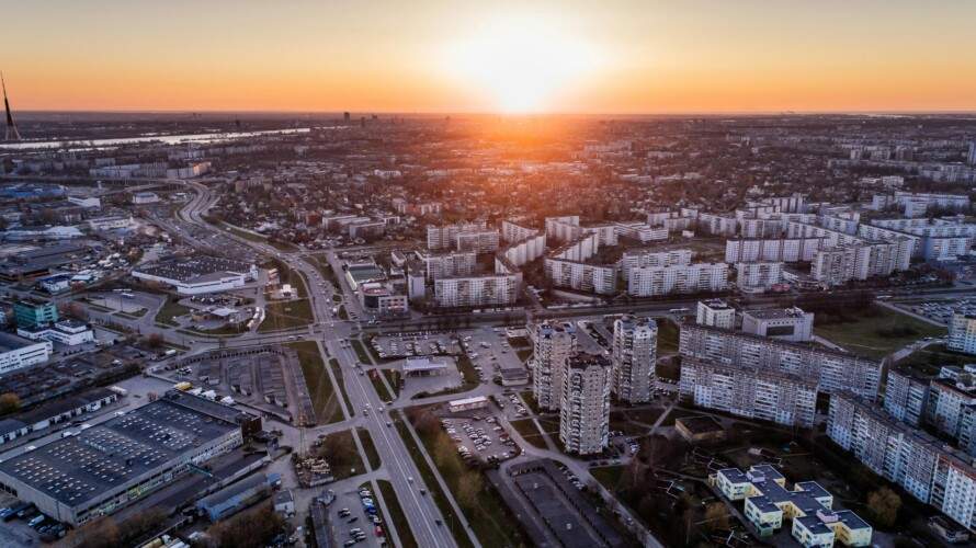 Aerial view of a cityscape during sunset, with residential buildings, roads, and parking lots in the foreground, and the sun setting on the horizon.