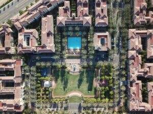 Aerial view of a large residential complex with a central grassy area, a swimming pool, palm trees, and surrounding multi-story buildings.