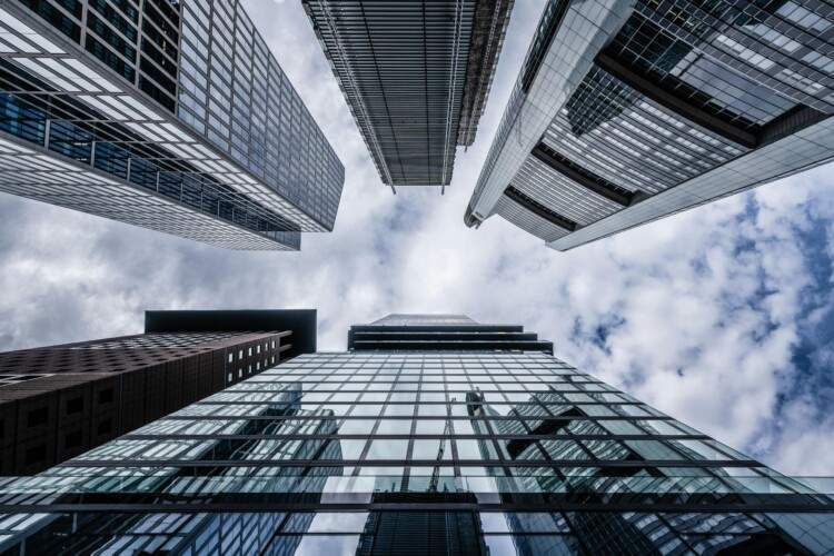 Looking up at several tall skyscrapers against a cloudy sky, with reflections on their glass surfaces.