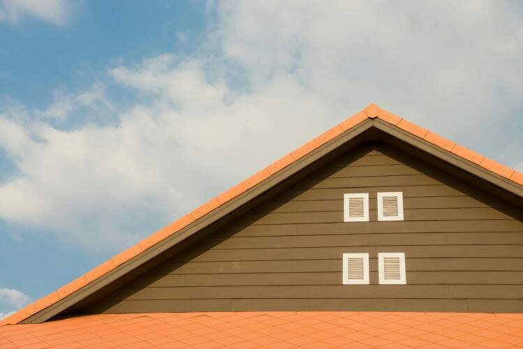 Roof of a house with orange tiles and brown siding, featuring two small white shuttered windows. Blue sky with clouds in the background.