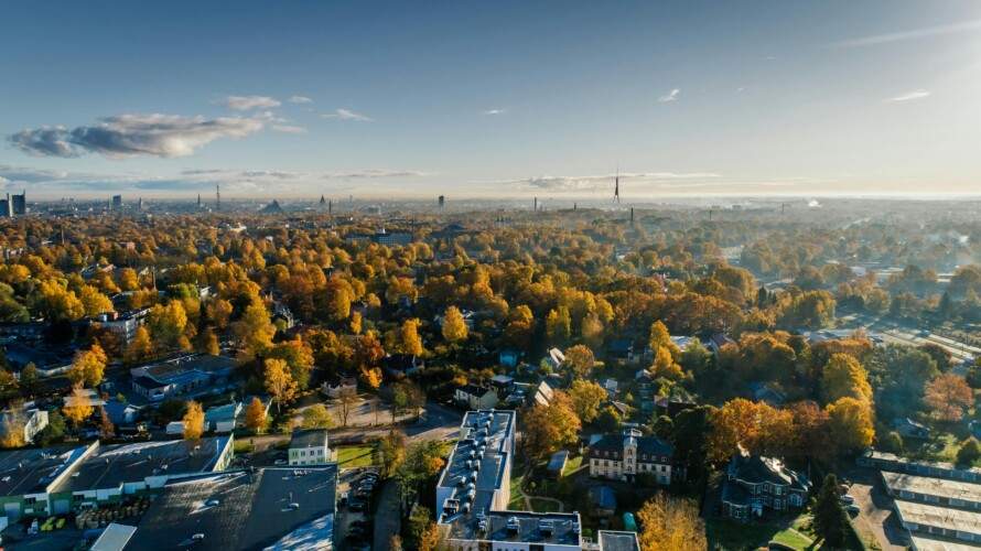 Aerial view of a cityscape with trees in autumn colors, buildings, and a clear sky in the background.